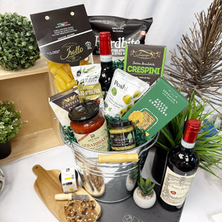 A gourmet gift basket filled with an assortment of Italian food products, including wine bottles, cheese, crackers, and pasta, arranged in a galvanized metal bucket with decorative plants around it.