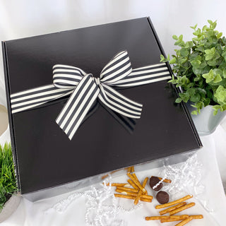 Black gift box with a striped bow on a white surface with snacks and plants.