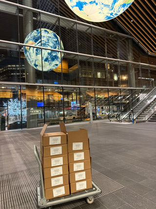 Stack of cardboard boxes containing corporate gifts on a cart in a modern building with large glass windows.
