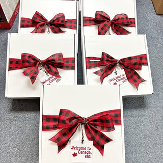 Gift boxes with red and black plaid bows and 'Welcome to Canada, eh!' tags on a gray surface.