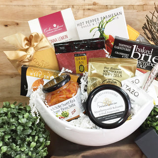 Gourmet gift basket with snacks, condiments, and a bottle of wine on a wooden surface.