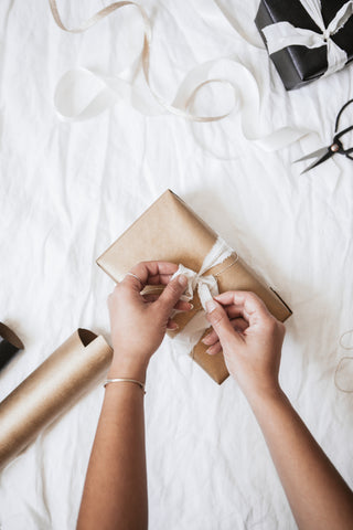 Person wrapping a gift with brown paper and white ribbon on a white surface.